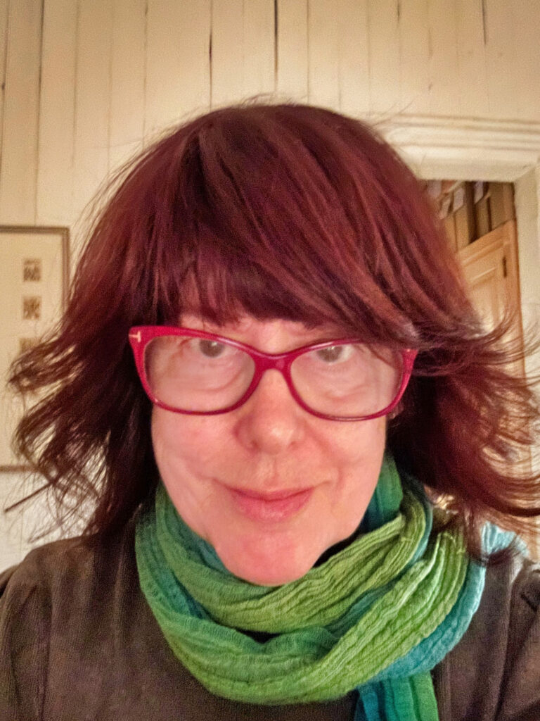 Headshot of Tricia Vita, with a green scarf around her neck and bright red glasses.