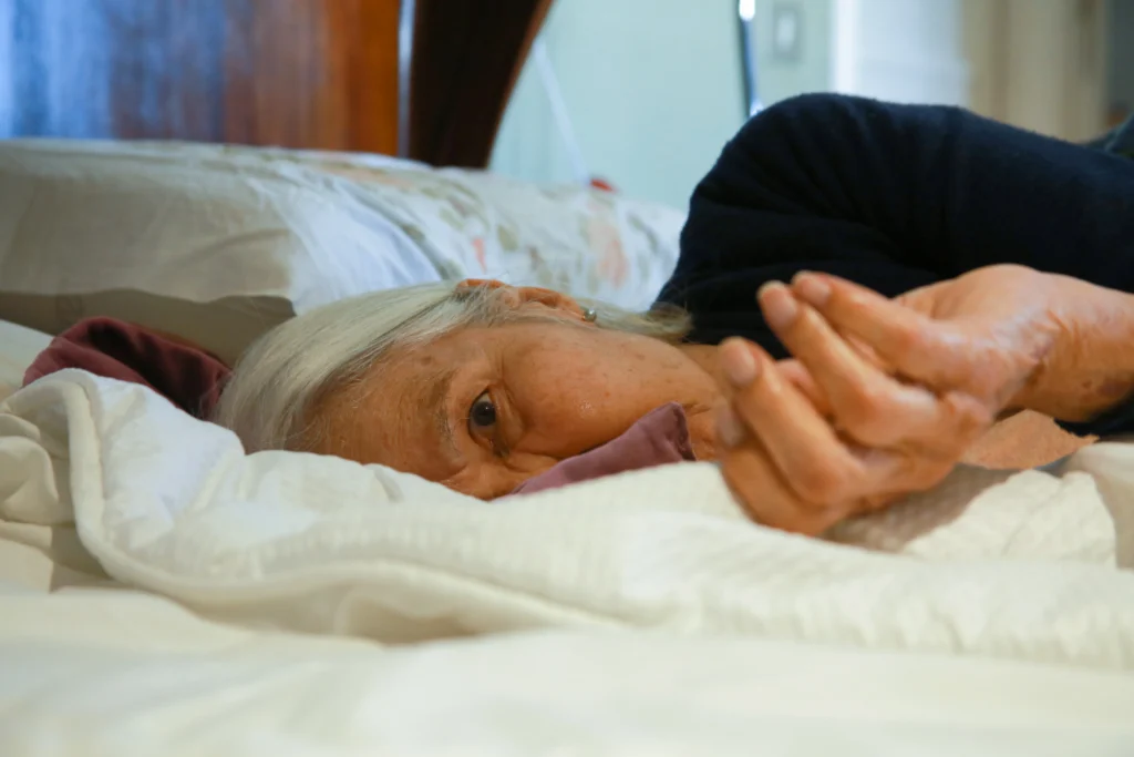 Image of Guadalupe laying on the bed, looking somber into the camera, the blankets slightly obscuring her face