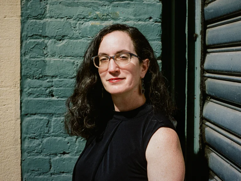 Woman in glasses, wavy brown hair, and a black shirt stands before a green brick wall.