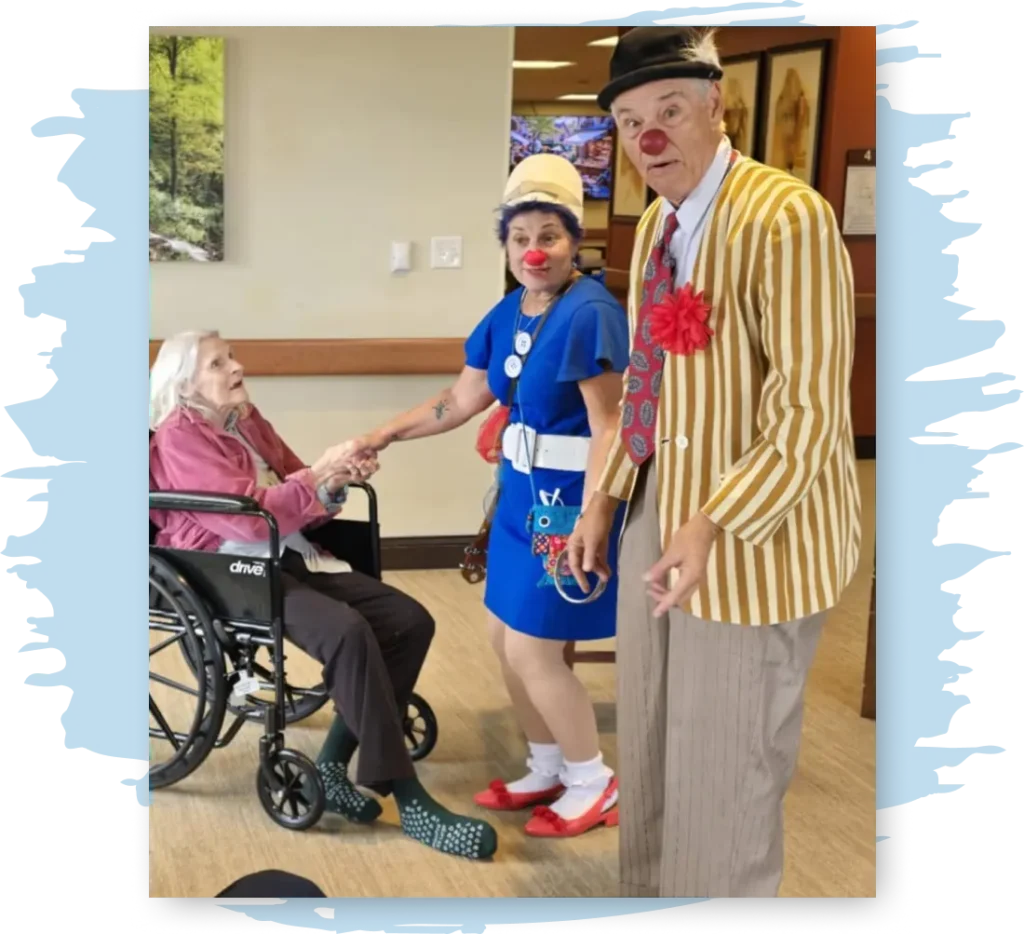 Two people dressed in a similar style to clowns joke and laugh with an elderly woman in a wheelchair