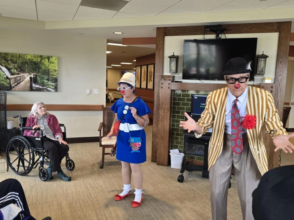 Two people dressed in playful outfits joke and engage a room of elderly individuals