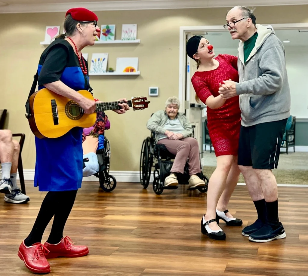 Woman dressed similar to a clown holds a guitar and sings with another couple dancing.