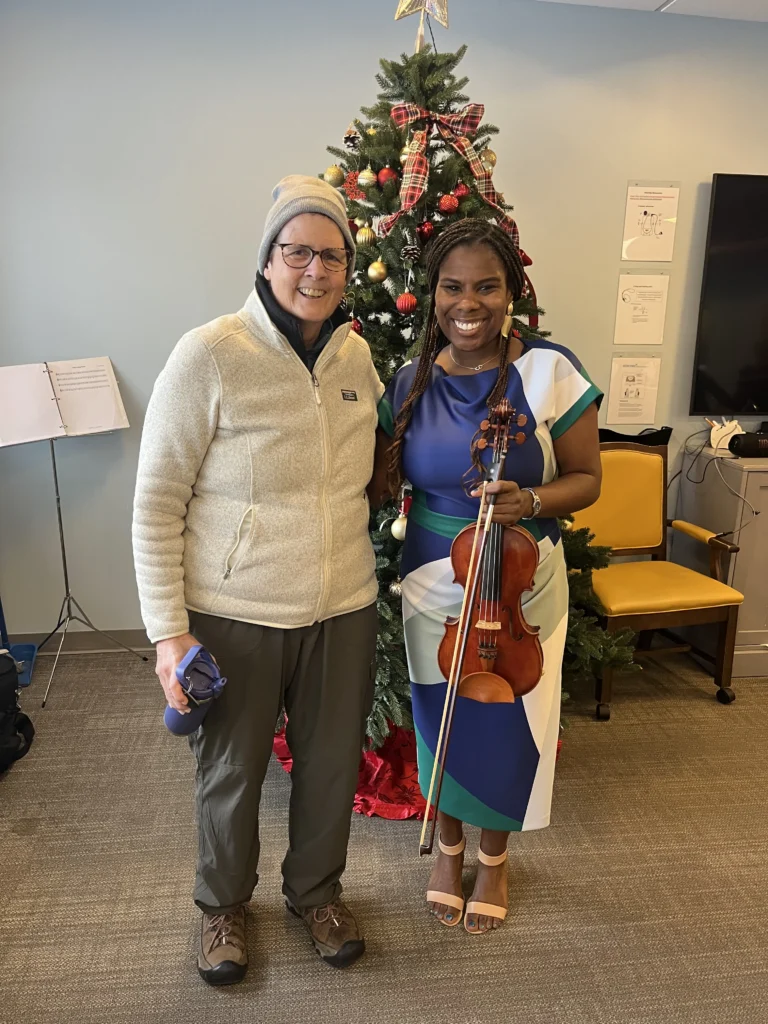 Melanie stand next to a staff member, while holding her violin, in front of a christmas tree