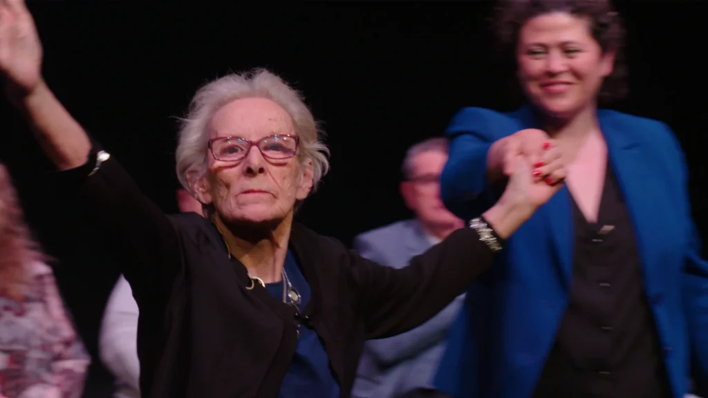 An older woman raises her hands at the audience after a perfomance