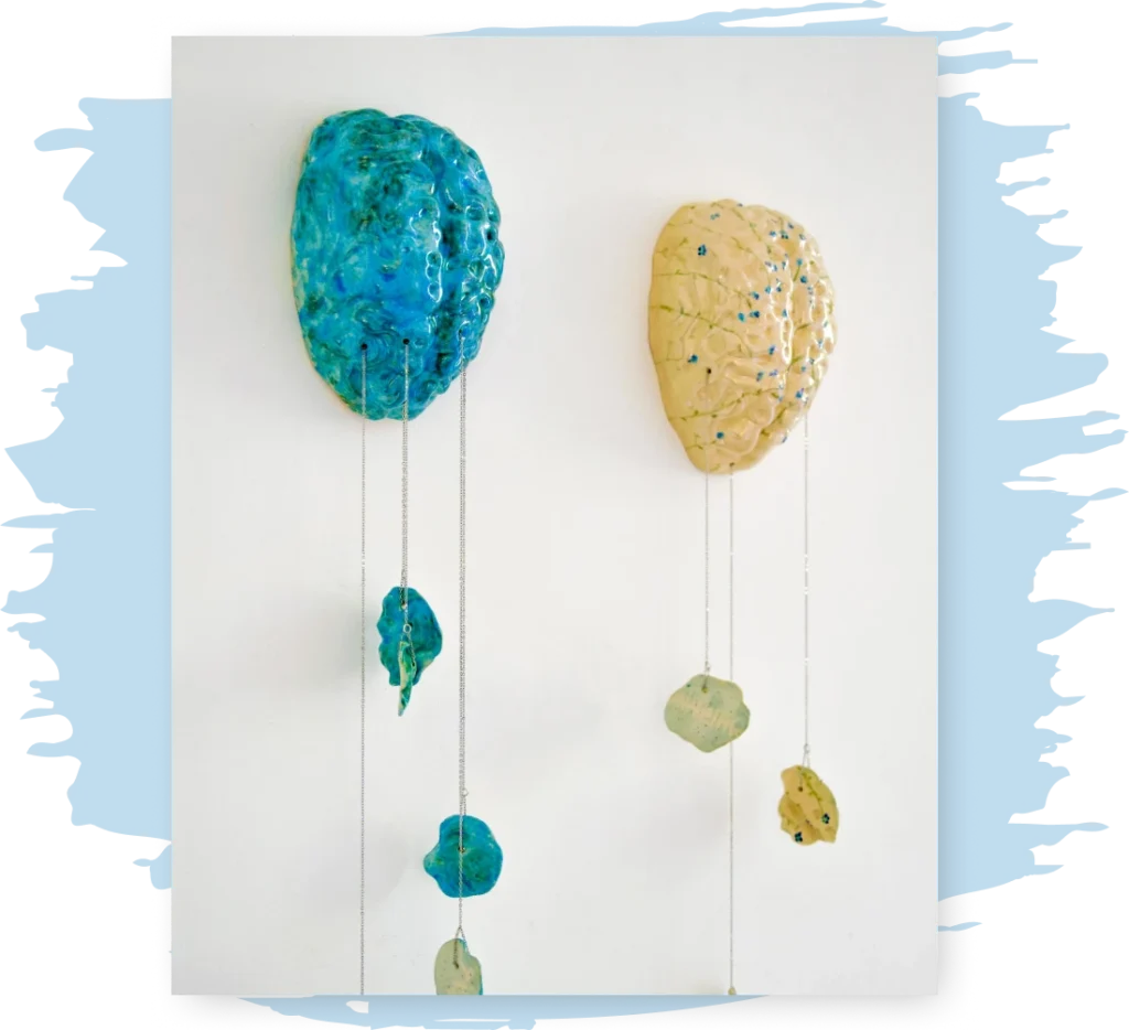 two ceramic brains hang from a wall, one blue one yellow, with pieces of the brain broken and hanging downward by chains.