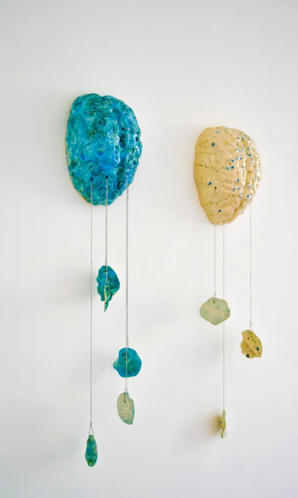 two ceramic brains hang from a wall, one blue one yellow, with pieces of the brain broken and hanging downward by chains.